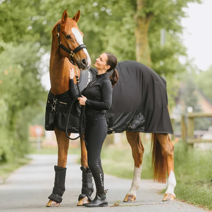A woman in black riding apparel stands beside a chestnut horse outdoors on a path, with the horse wearing a black turnout blanket and black leg wraps. Trees and a blurred rural background are visible.