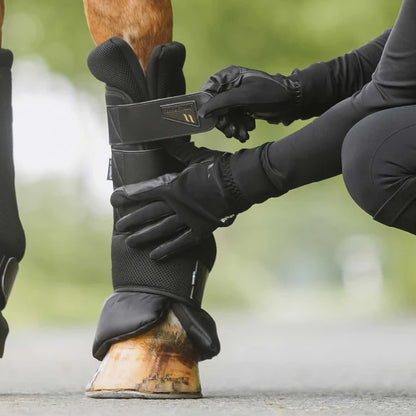 A close-up of a horse leg being fitted with a black airflow quick wraps by a person wearing black gloves, with a blurred outdoor background.