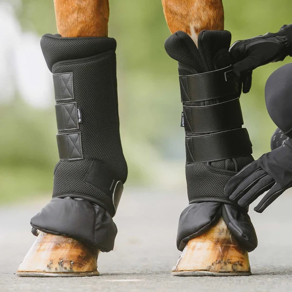 A close-up of a horse leg being fitted with a black airflow quick wraps by a person wearing black gloves, with a blurred outdoor background.
