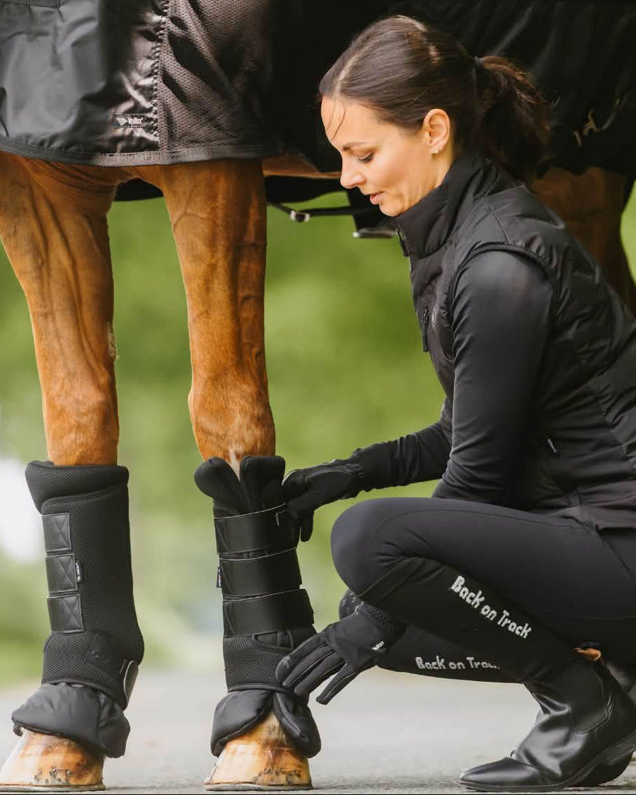 A woman in black equestrian apparel crouches beside a horse wearing black airflow quick wraps and a black mesh rug.